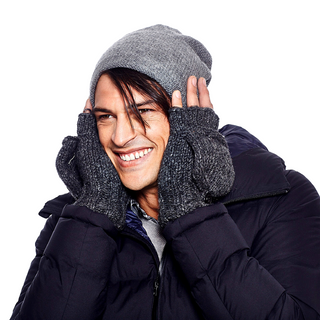 Dressed in winter attire, a person smiles warmly while wearing a grey knit beanie, dark blue puffer jacket, and dark grey Fingerless Gloves With Button Flap and Fleece Lining. Holding their cheeks with their gloved hands, they radiate cheerfulness against the white backdrop.