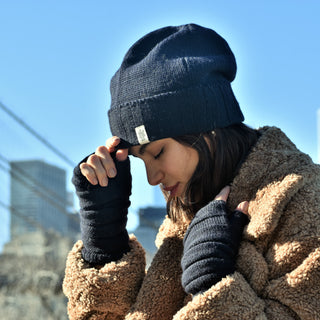 A person dressed in a brown teddy coat and a black knit hat stands outdoors against a backdrop of urban buildings and a blue sky. The individual, showcasing impeccable winter style with Reverse Step Handwarmers, looks down with one hand adjusting the hat as sunlight illuminates their face.