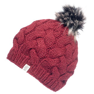 The Boheme Cable Beanie w/ Faux Fur Pom is a knitted red winter hat featuring a thick cable pattern and a faux fur pom-pom on top. Made from 100% Merino Wool, it includes a small, white tag with a logo near the edge of the brim.