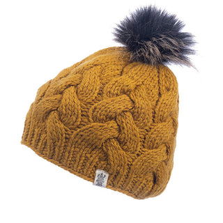The Boheme Cable Beanie w/ Faux Fur Pom is a mustard-colored, cable-knit winter beanie made of 100% Merino Wool, featuring a black and brown faux fur pom-pom on top. It has a label with a logo at the bottom edge.
