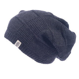 The Checkered Slouch, a dark gray knitted beanie with a checkered pattern, is displayed against a white background. Handmade in Nepal, it boasts a textured design and features a small, light-colored tag with an emblem sewn near the edge.