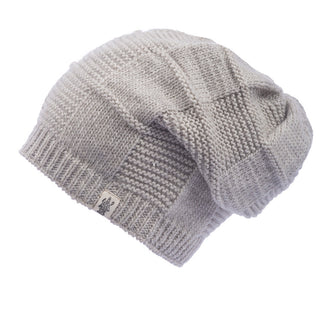The Checkered Slouch is displayed against a white background, featuring a gray knitted beanie with a checkered pattern. The beanie has a folded brim, a small fabric tag with a logo on it, and boasts a chunky texture. This wool hat is handmade in Nepal.