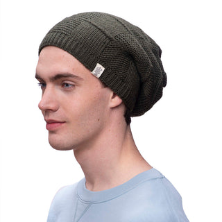 Profile view of a person with light skin and short brown hair wearing a dark green knit Checkered Slouch beanie handmade in Nepal and a light blue sweatshirt. The beanie, adorned with a small logo tag on the side, pairs stylishly against the plain white background.