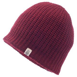 The Cardigan Knit Beanie is a red, handmade accessory from Nepal, featuring a textured knit pattern. It’s crafted from warm, 100% wool material and has a small white tag with a logo sewn onto the lower edge.