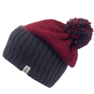 The Masha Pom Slouch is a handmade, knitted beanie featuring a dark gray ribbed fold-over cuff and a solid red top. It is adorned with a multicolored pom-pom in shades of red and gray and has a small fabric tag with the logo attached to the cuff. Crafted from cozy wool, this hat showcases its unique design perfectly.
