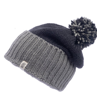 The Masha Pom Slouch is a handmade knit beanie featuring a bottom ribbed section in light gray and a top section in dark gray. It includes a pom-pom on top, crafted from both light and dark gray wool yarn, and sports a small tag with a logo on the ribbed section.