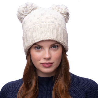A person wearing a cream-colored Double Pom Kira Hat made of wool, paired with a navy blue sweater, against a plain white background.