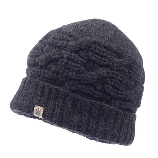 The Side Cable Knit Beanie is a black cable knit hat featuring a ribbed fold-over brim and a small beige tag with a logo. Handmade in Nepal from 100% wool, this beanie boasts a textured pattern and appears to be thick and warm.