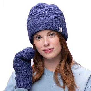 A person wearing a blue Side Cable Knit Beanie, handmade in Nepal, along with matching mittens and a light blue sweatshirt. The individual has long red hair and is standing against a plain white background.