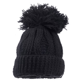 The Big Pom Rib Fold Hat is a black, knitted winter beanie featuring a folded brim and a large pom-pom on top. It boasts a cable knit pattern and is handmade in Nepal from 100% merino wool.