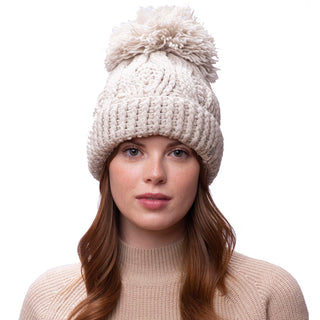 A person with long, wavy hair wears a Big Pom Rib Fold Hat, made from 100% merino wool and handmade in Nepal. The hat features a large pom-pom on top. They pair it with a cozy beige sweater and gaze directly at the camera against a plain white background.
