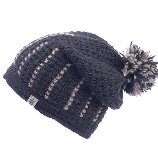 The Ferry Slouch Hat is a gray knitted beanie made from luxurious merino wool, featuring cream-colored vertical stripes and a matching pom-pom on top. Handmade in Nepal, it includes a small tag with a logo on the side for added charm.