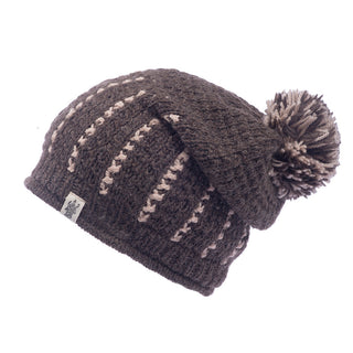 The Ferry Slouch Hat, a delightful brown knitted beanie crafted from soft merino wool, is highlighted with cream-colored stripes and a charming pompom on top. Handmade in Nepal, it includes a small label on the side and is displayed against a simple white background.