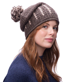 A person with long red hair wearing the Ferry slouch Hat, a knitted brown beanie crafted from merino wool, and a blue sweater, looking to the side against a plain white background.