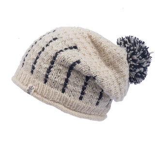 The Ferry slouch Hat is a handmade, cream-colored knitted beanie crafted from soft merino wool, featuring dark vertical stripes and a playful pom-pom on top, bringing a touch of Nepalese charm.