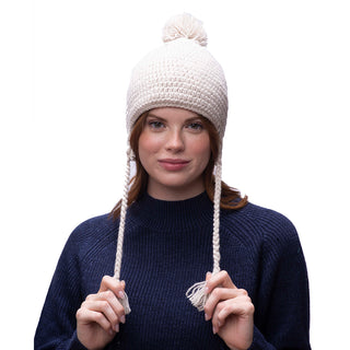 A person wearing a navy blue sweater stands against a plain white background, with the Verona Earflap Hat in cream adorned with wool earflaps on their head. The hat, designed for extra warmth with its sherpa lining, features braided tassels that they hold in their hands, all while maintaining a relaxed expression.
