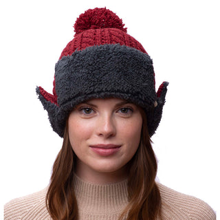 A person wearing the Sherpa Trapper Hat w/ Pom, which showcases red and gray colors. Handmade in Nepal, this hat features a pom-pom on top and ear flaps designed to resemble elf ears. The person has long hair and is dressed in a beige sweater.