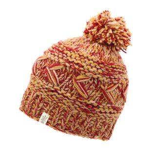 The Reverse Cross Pom Pom Hat features an intricate knitted pattern in shades of red, orange, and pink. It is topped with a large pom-pom and has a small white tag with a logo on the side. The texture appears thick and warm.