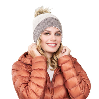 A woman wearing a brown puffer jacket and the Stripe Flower Fur Pom Pom Merino wool hat smiles while slightly adjusting her hat, against a plain white background. Her blonde, wavy hair is visible under the winter accessory.