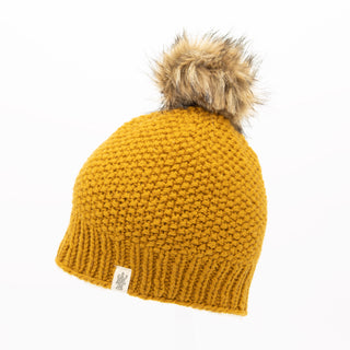 The Moss Fur Pom Pom Hat is a mustard yellow knitted beanie featuring a ribbed brim and a faux fur pom-pom on top. It includes a small fabric label with a logo near the brim, set against a plain white background.