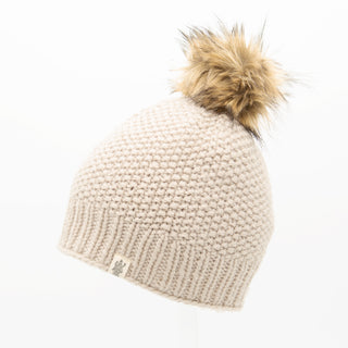 The Moss Fur Pom Pom Hat is a beige knitted beanie featuring a faux fur pom-pom on top. It has a ribbed brim and a small tag with the logo near the base, all set against a white background.