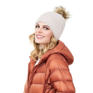 A person with long, wavy blonde hair is wearing the Moss Fur Pom Pom Hat and an orange puffer jacket. The person is smiling and looking to the side against a plain white background.