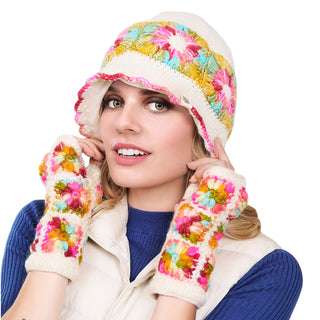 A person is wearing a hand-knit Mushroom Hat and fingerless gloves, both adorned with colorful flower patterns. They are also dressed in a blue top and a light vest. The person is touching the sides of the hat and looking slightly upward against a white background.