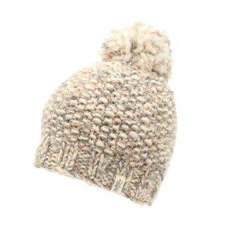 A product shot of the Chunky Pom, a beige and gray knitted beanie hat adorned with a pom-pom on top. It showcases a textured, chunky knit pattern and includes a small branded tag on its ribbed brim. The background is completely white.