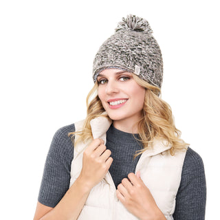 A smiling person with long, wavy blonde hair is wearing the Chunky Pom gray beanie and a white puffer vest over a gray ribbed top. The background is plain white.
