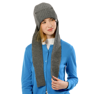 A person sporting a blue cardigan and the grey Pelham Hat made of merino wool, featuring long ear flaps. They are smiling slightly and holding one of the hat's ear flaps with their right hand against a plain black background.