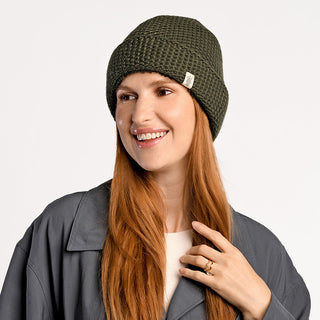 A woman with long red hair wears the Baltic Hat, an olive green wool hat handmade in Nepal, along with a gray jacket over a white top. She smiles, looks slightly left, touches her hair with one hand, and wears a ring.