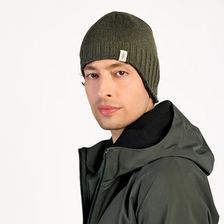 Someone wearing the Dakota Hat, an olive green wool beanie, and a dark green hooded jacket looks at the camera against a plain white background.