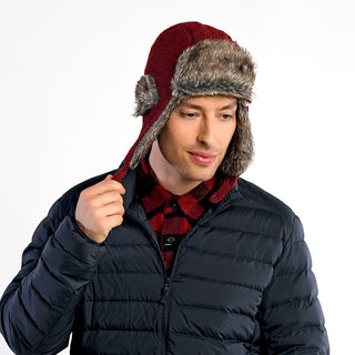 A man in a dark puffer jacket wears the Fern Hat, a red winter trapper with faux fur trim, and holds one ear flap while looking down. The background is plain white.