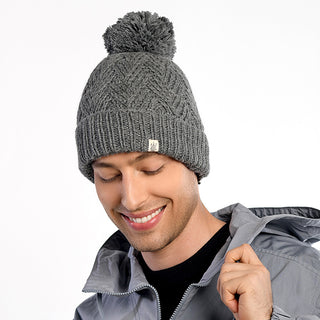 A man wearing a gray Aspen Hat with a pom-pom, along with a gray jacket and black shirt, smiles while looking down against a plain white background.