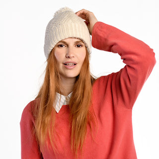 A woman with long red hair wears the Aspen Hat, a cream-colored wool winter accessory, paired with a coral sweater over a white collared shirt. She touches the top of her hat and gazes to the side against a white background.
