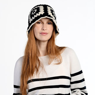 A woman with long red hair wears the Milie Hat, a crochet wool beanie with a geometric pattern, and a cream sweater with bold black stripes, standing against a plain white background.