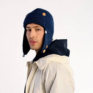 A man wearing a cream-colored jacket, a black hoodie, and the handmade Elk Hat—an outdoorsy wool hat from Nepal with ear flaps and wooden buttons—stands against a plain white background.