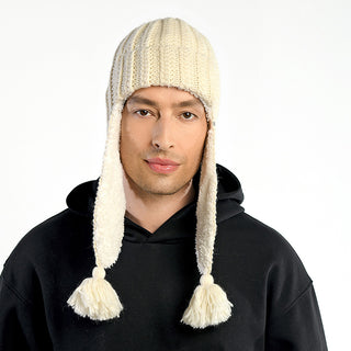 A man wears a cream-colored Alex Hat, a wool winter hat with long ear flaps and tassels handmade in Nepal, paired with a black hoodie, standing against a plain white background.