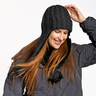 A person with long reddish-brown hair smiles while adjusting the Alex Hat, a black faux shearling style, and wears a dark puffy jacket against a plain white background.