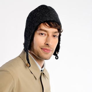 A man wears the Doris Hat, a dark gray wool style with faux shearling lining, paired with a beige coat, as he looks at the camera against a plain white background.