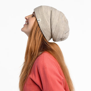 A person with long reddish-brown hair wears the Edgar Slouch beanie and a coral pink top, smiling in profile with eyes closed against a plain white background.