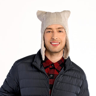 A man stands against a plain white background wearing the Ezra Hat, a handmade beige wool hat with ear flaps and pointed corners, along with a red and black plaid shirt and a dark puffer jacket.