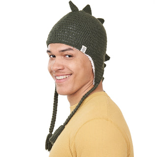 A person wearing a DinoTail Hat, which is green and crocheted with spikes and ear flaps, is smiling. The hat features braided ties hanging down, and the individual is dressed in a yellow, long-sleeved shirt. They are turned slightly to the left with their head turned towards the camera.