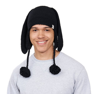 A person is wearing the Long Ear Puppy Hat, which features long ear flaps and pom-pom tassels. They are smiling in a light gray shirt against a plain white background.