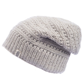 The Boho Slouch, a light gray knitted beanie perfect for winter, is displayed against a plain white background. This beanie features a folded brim and a label with a small emblem on the side. Its knit pattern is textured with a mix of tight and loose stitches, giving it a cozy appearance.