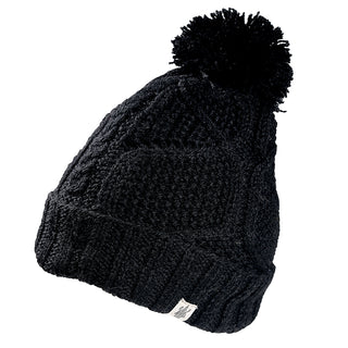 The Cedar Hat is a black knitted beanie with a folded brim, a pom-pom on top, and a small white fabric tag on the cuff.