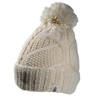 The Cedar Hat is a cream-colored knitted beanie with a thick ribbed edge, cable knit pattern, large pom-pom on top, and a small fabric tag on the brim. Shown against a white background.