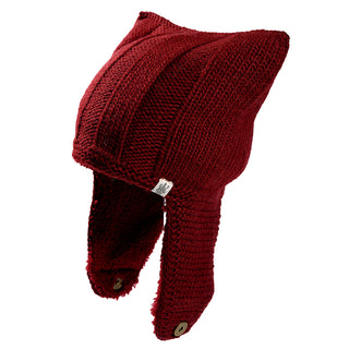 The Ezra Hat is a red knitted hat with two pointed cat ear tips, ear flaps that fasten under the chin with buttons, and a small white tag on one flap.