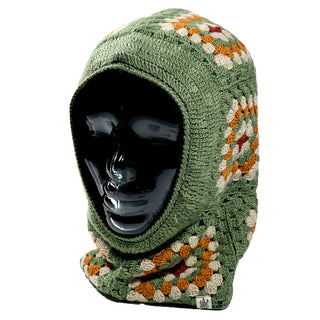 A glossy black mannequin head displays the Ryan Hood, a green crocheted balaclava featuring orange, white, and brown squares. The Ryan Hood covers the entire head and neck while leaving the face exposed.