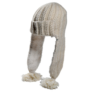 The Alex Hat is a cream-colored knit winter hat with ear flaps lined in soft material and large pom-poms at the ends, shown on a white background.